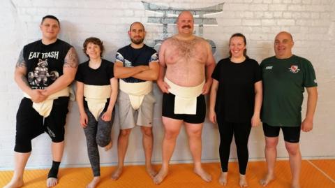 The image shows six people standing side by side in a martial‑arts or sumo‑training space. They are positioned on an orange padded floor in front of a white brick wall. Most are wearing a white mawashi over their clothing.