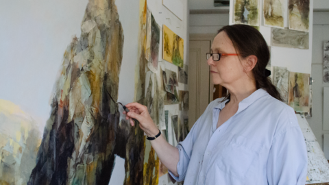 A woman with long brown hair and glasses paints with oils on a large canvas.