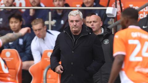 Blackpool manager Steve Bruce staring out over the pitch. He has grey hair, and is wearing a black jacket. In front of him are Blackpool players wearing their Tangerine coloured shirts.