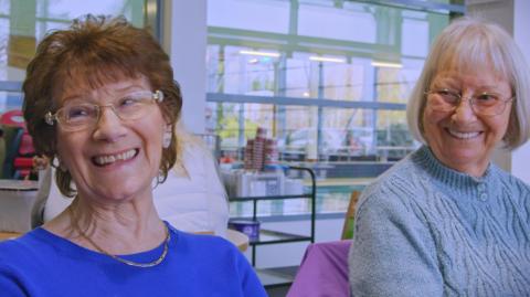 Two women smiling in a leisure centre.The woman on the left is looking away from the camera, wearing glasses, a gold necklace and a blue jumper. The other day is hearing a light blue jumper, has glasses on, and has bobbed, fair hair. She is smiling in the direction of the other woman. Behind them is pool and leisure equipment. 