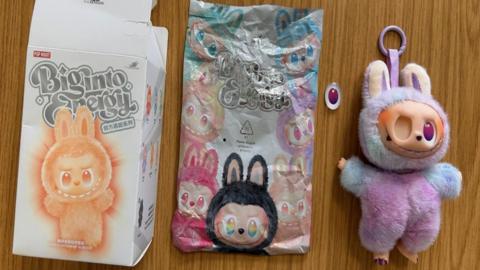 White cardboard packaging and paper packaging alongside a doll with its eye detached.