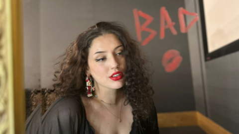 The image shows a woman with curly dark hair and red lipstick, dressed in a black top and wearing large earrings, gazing slightly off to the side. It looks as if the word "BAD" is boldly written in red lipstick on a mirror in front of her and to the side, with a kiss mark. 