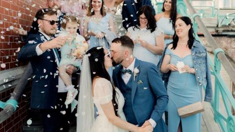 Ally and Paul kissing while wedding guests throw confetti over them in celebration 