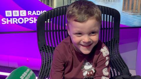 A boy in a radio studio. He is wearing a purple top with a Mickey Mouse logo