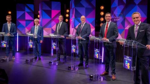Six men in business suits lined up behind podiums smiling at the camera in a television studio 