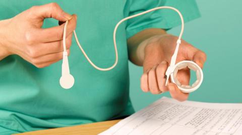 A man in green medical clothing holds a small padded ring in one hand, linked with a thin cable to another white device