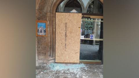 An arched stone entrance to a church which has been partially covered up by a wooden board. Beneath the board are many fragments of light blue shattered glass. To the right of the board is a second automatic door which is still intact.