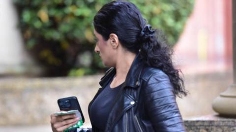 A woman with long dark hair and wearing a dark leather jacket walking outside court - she has her head turned away from the camera and a phone in her hands
