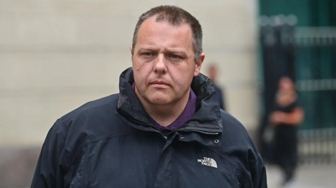 A man wearing a black North Face jacket and a purple shirt. The background of the image is blurred. 