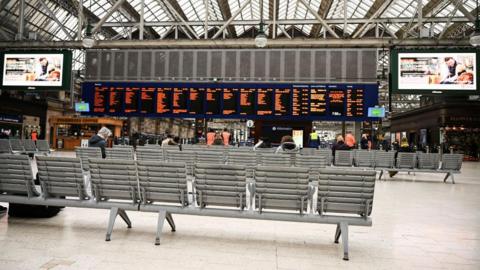 People sit in metal sits in the main concourse of Glasgow Central Station, looking at black and yellow departure boards. 