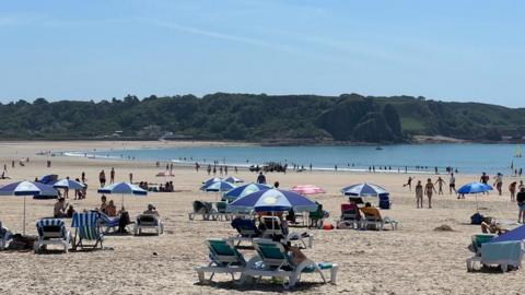 A busy beach on a summer day shows several blue and white sun-loungers and beach umbrellas. The tide is out, with many people in the shallow waves. There are also several sunbathers on the sand. In the background, green hills and trees rise above the bay.