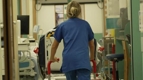 An NHS worker pushing a bed through a hallway in a hospital. She is wearing blue scrubs and has her blonde hair up in a bun. 