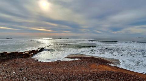 A pebbly Lyme Regis beach sitting in front of a choppy looking sea under cloud that is coloured by tints of orange from a low sun 