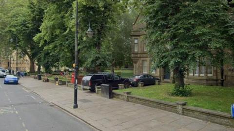 An empty taxi rank against the backdrop of a lamp-post, trees and a large stone building.