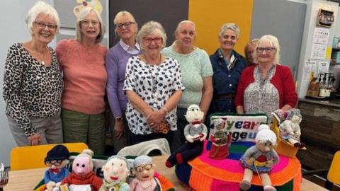 Eight women are standing in a line in front of a table, smiling. There are two postbox toppers on the table with knitted figurines on them. Some are knitting, some are sitting down and one is on a chair. one of the toppers reads "75 years age uk"