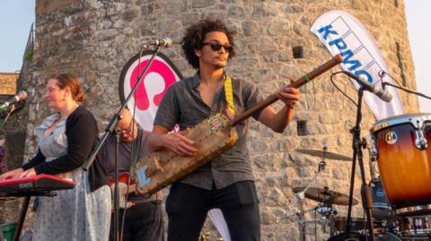 A man wearing sunglasses, a grey shirt and black trousers stands in the centre of the image on stage in front of a castle wall. He is playing an instrument that looks similar to a guitar. Behind him is a woman playing the electric keyboard and a man playing the electric guitar. To the right of the image is a drum kit.