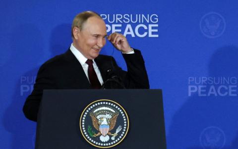 Putin stands at behind a podium, smiling. He is wearing a dark suit and holding an earpiece