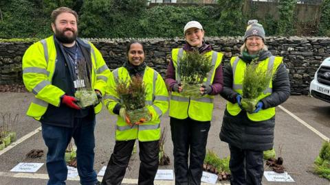 A man and three women in high-viz coats hold saplings in their hands, ready to hand them out to the public. They are standing in a car park and they are all smiling at the camera.