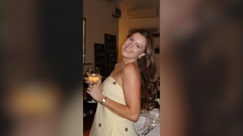 A young woman with long brown hair smiles while holding a glass of wine. She is wearing a strapless yellow dress.