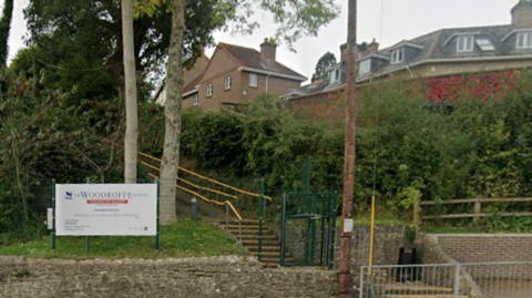 Woodroffe School buildings are partially visible above a hedge as seen from the road. A white school sign is at street level.