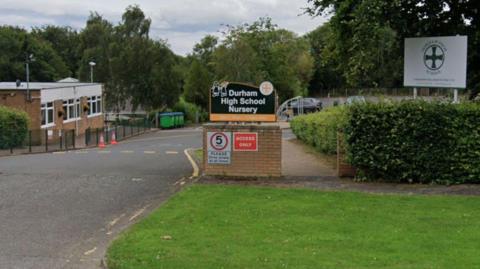 View of the entrance to Durham High School and Nursery, with school signs, at the head of an access road, hedges on the perimeter and school buildings visible in the background.