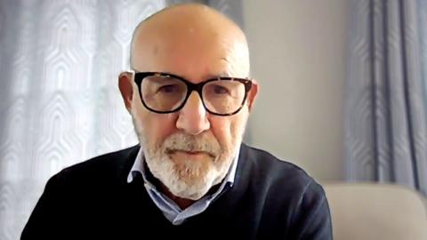 Jon Trickett is in a blue shirt, black jumper and black glasses. He is a bald man with a grey goatee