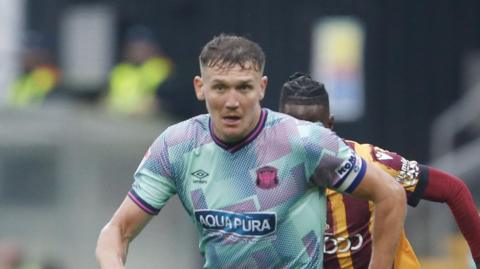 Charlie Wyke playing for Carlisle in September 2024 away to Bradford City with a Bradford player right behind him with his face obscured 