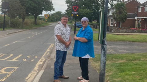 Two people looking wards camera standing in front of a Vehicle Activated Sign