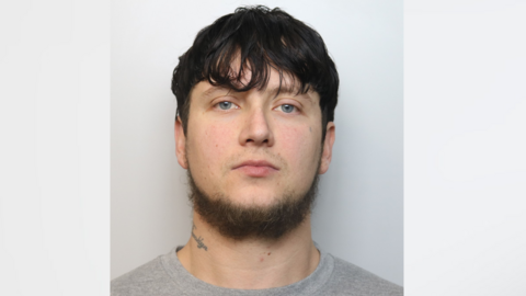 A man with black hair, a slight beard and a neck tattoo. He looks seriously at the camera in a police photo.