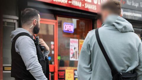 A GMP officer in a grey hoodie with a black police vest holds his walkie talkie stood next to a man in a teal hoodie with his face blurred. They are standing in front of a kebab shop. 