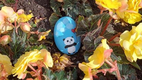 A rock coloured blue with a panda holding heart-shaped balloons. It is in a flowerbed.