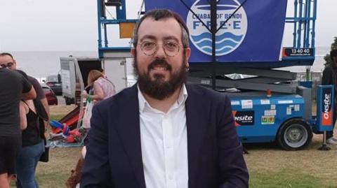 A mid shot of Rabbi Eli Schangler standing in front of a vehicle which has the branding of Chabad on. He has short dark hair, a long black beard and is wearing a white shirt and dark jacket.