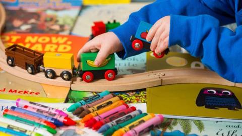 Close up of a young child playing - showing their hands and arms only. They are holding a two sections of a wooden train set, with two other sections running on wooden rails. To the far right part of a book can be seen with a front cover depicting a cartoon blue and brown owl on a dark yellow background. Other books are in the background, slightly out of focus. In the foreground are lined up about a dozen large and brightly coloured crayons.