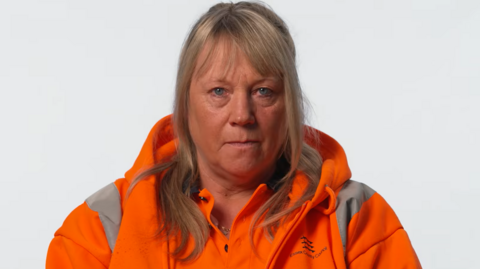 A recycling centre employee sitting in front of a white background. She has long blonde hair and is staring at the camera with a serious expression on her face. She is wearing an orange polo top with a hi-vis orange jacket over the top.