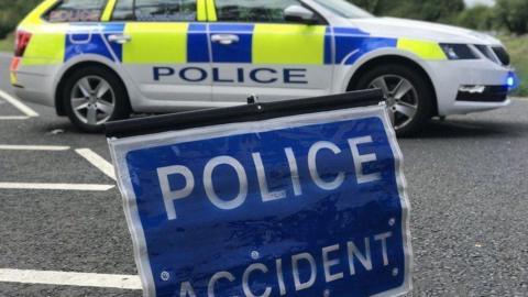 A blue sign saying police accident in white capital letters. It is sitting on a tarmacked road. Behind it is a white, blue and yellow police car and behind that, a verge. 