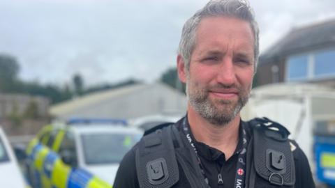 A bearded police officer is wearing a protective vest. There are several Devon and Cornwall Police vehicles in the background. They have fluorescent yellow and blue markings.