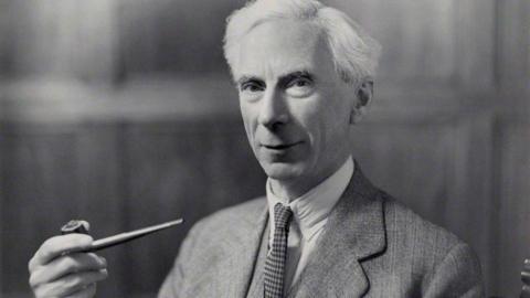 A black and white picture of Bertrand Russell looking at the camera. He has short white hair and is wearing a shirt, a tie and a jacket. He is holding a pipe in one hand.