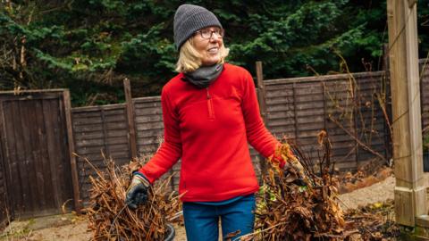 A woman in a wooly hat, gloves and scarf carries two buckets of garden waste.
