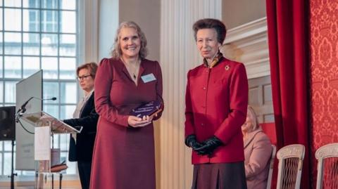 A woman looking at the camera in a red dress holding an award. She is smiling and stoop next to Princess Anne who is wearing a red jacket and black gloves.