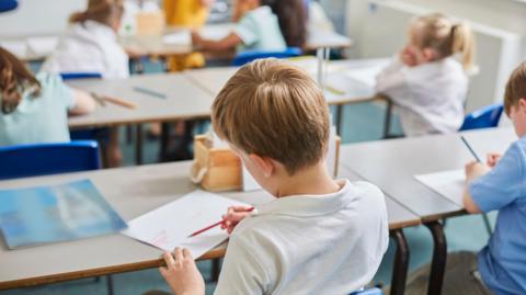 A young school boy wearing a white polo shirt is sat in a classroom looking away from the camera doing work with a red pencil on a white sheet of paper.