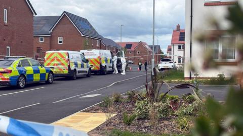 Two officers wearing all white forensics clothing stand at the open door of a police van. It is parked at the side of a road on a residential estate, ahead of two other police vehicles. Blue and white police tape can be seen in the foreground and further down the road in the background.