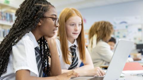 Two teenage girl high school students studying in front of a laptop in a schoolroom. The pupil on the right has long black curly hair and dark-framed glasses. The one on the right has long red-blonde hair. A teacher and other pupils are seen blurred in the background