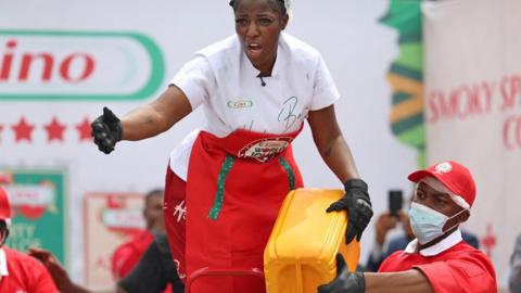 igerian chef Hilda Baci, dressed in a white shirt, red apron and black gloves, shouts and gestures as she holds a yellow carton together with a male colleague dressed in a red shirt, red cap and facemask