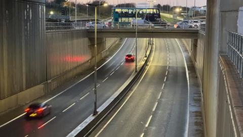 A main road seen from a traffic bridge above. Two cars travel along in one direction and cars and a bus move along another bridge over it. 