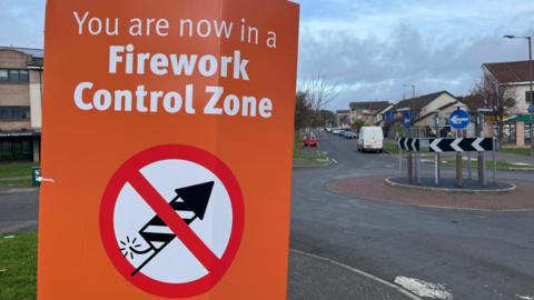 An orange sign warning people they are now in a firework control zone. The text on the sign is in white. There is a logo of a black and white firework on a white background with a red circle around it and a red strike through the middle.