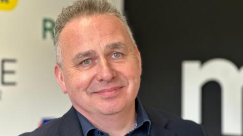 Matthew Marriott pictured smiling. He has short but spiked grey hair, wears a blue blazer and blue collar shirt.