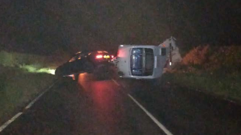 A caravan lying on its side beside an abandoned car with its lights on