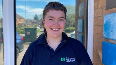 Isla is wearing a blue polo shirt with Stratford-on-Avon Foodbank written on the left hand side of the shirt. She has short hair and is smiling at the camera.