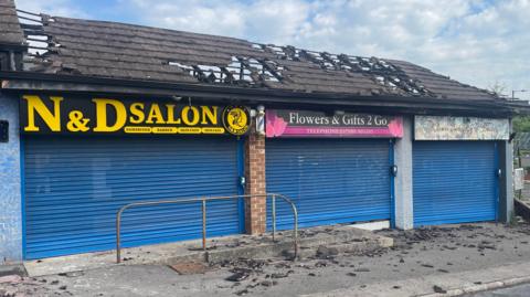 Front view of the shows, all with shutters down and the roof damaged. In places it has almost entirely fallen through.