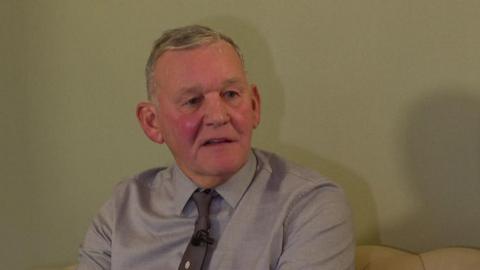 Keith Franks wearing a grey shirt and tie, in front of a grey background.